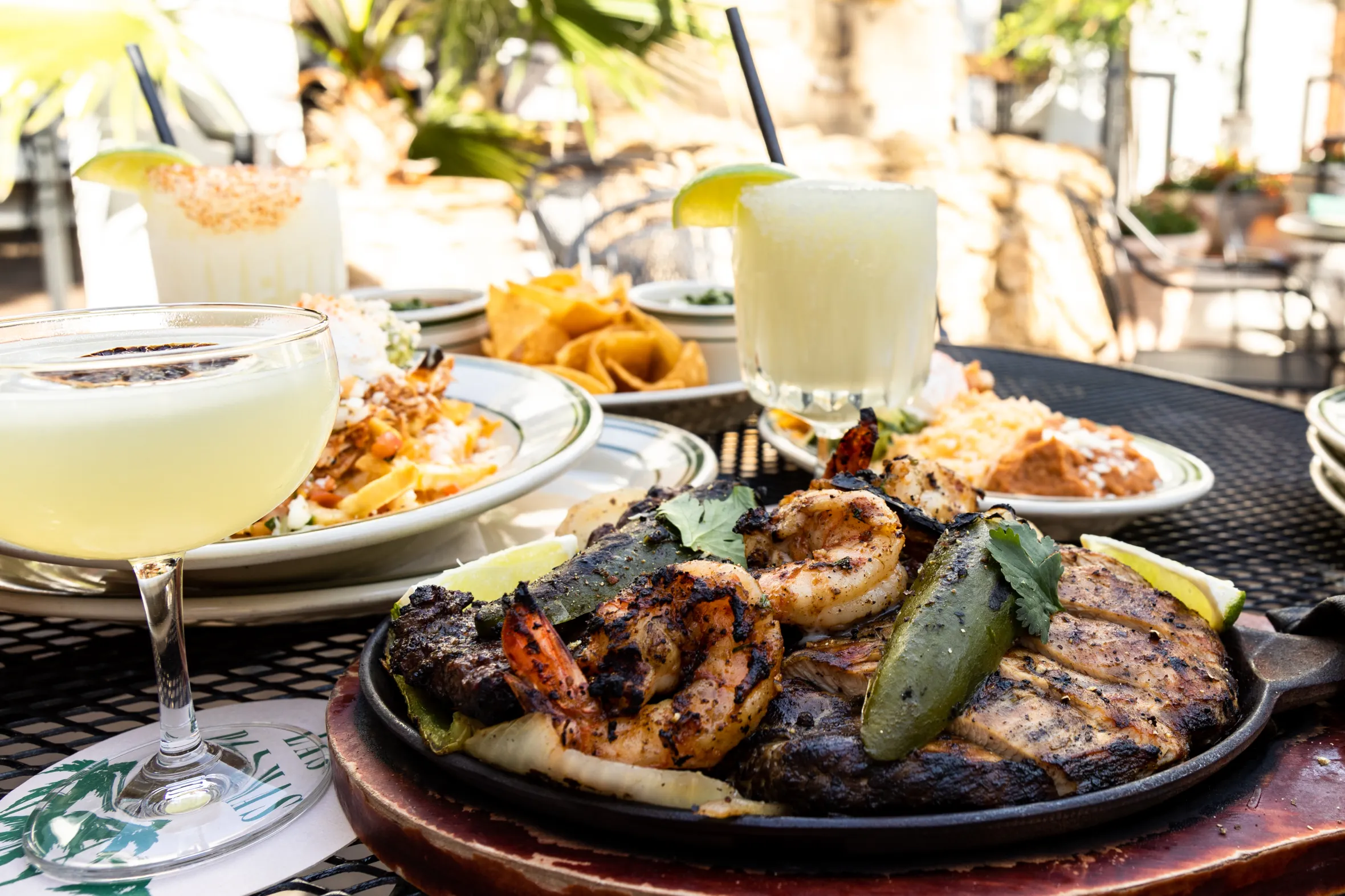 A table of various Mexican dishes and drinks, including fajitas with shrimp and chicken, nachos, and margaritas, on an outdoor patio.
