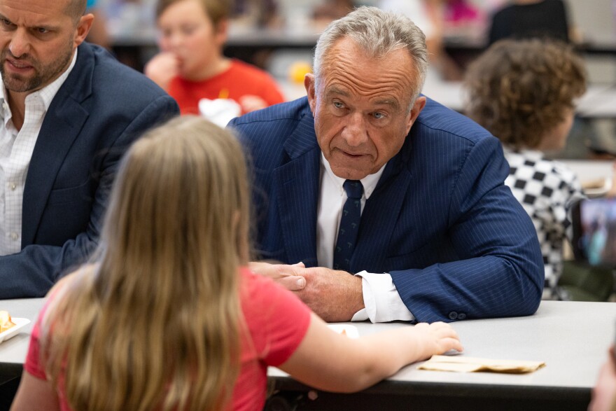A man in a suit sits at a cafeteria table talking with a young girl. 