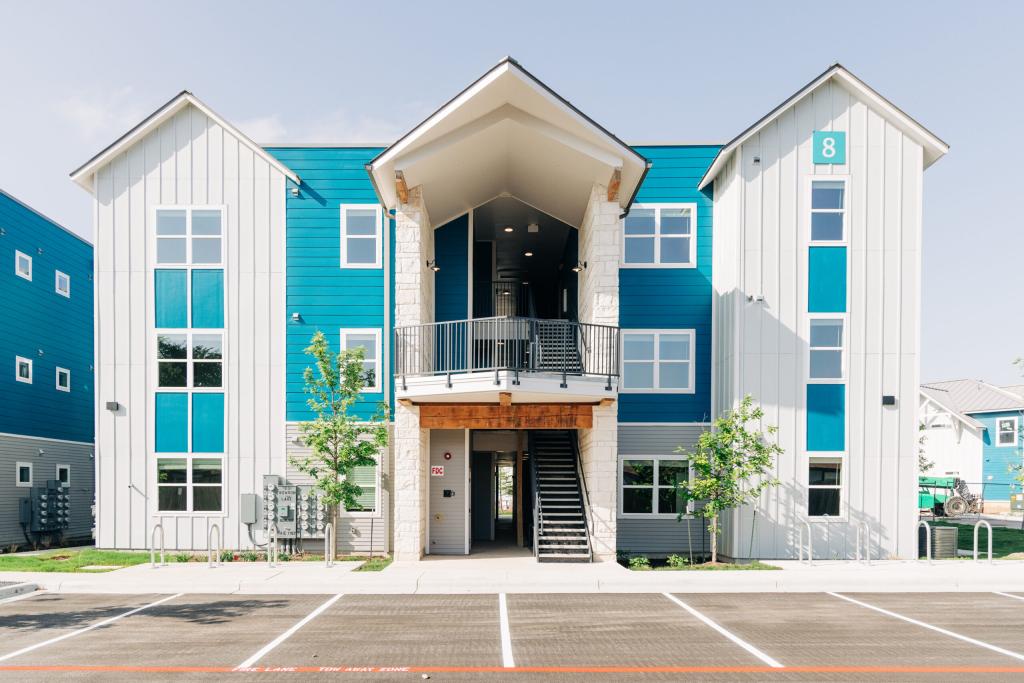 Image shows front-facing exterior view of one of the buildings at Persimmon Point. The exterior of the multi-story building features white vertical vinyl siding with blue horizontal siding. 