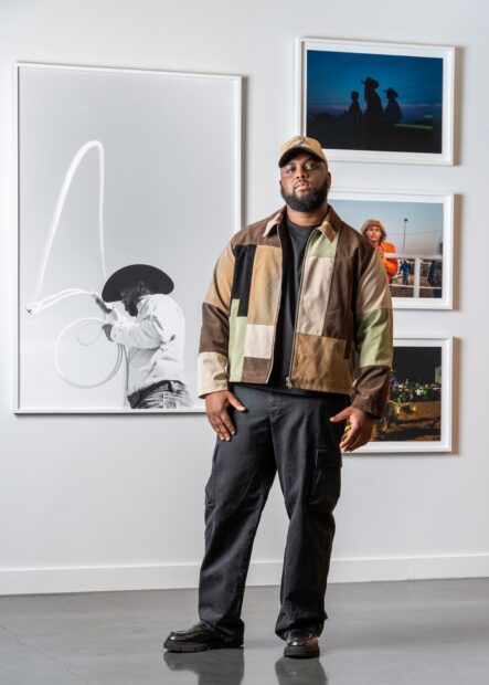 Artist Brian Edwards Jr. stands with several of his photographic artworks on a wall behind him.