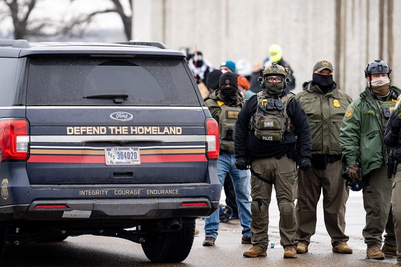 Masked ICE agents stand on the streets of Minneapolis next to a U.S. government SUV with "Defend The Homeland" emblazoned on it.