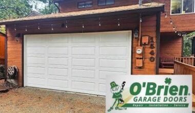 This image features a traditional white sectional garage door installed on a two-story home with rustic orange-brown wood siding. A decorative string of outdoor lights hangs above the door, while the foreground displays the official "O