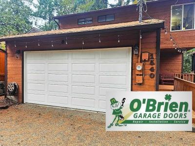 This image features a traditional white sectional garage door installed on a two-story home with rustic orange-brown wood siding. A decorative string of outdoor lights hangs above the door, while the foreground displays the official "O