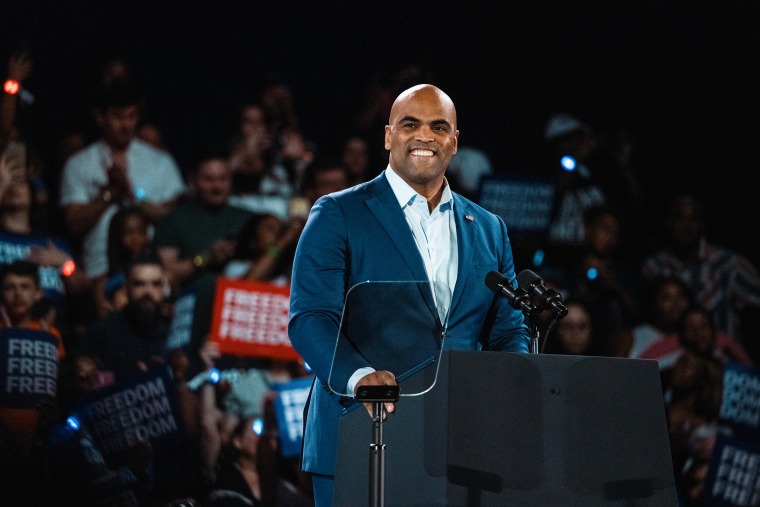 Colin Allred grins while standing at a lectern during an indoor rally.