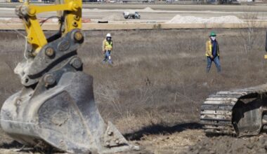 The bottom of a yellow excavator stand in the foreground with two construction works walking through an open site in neon yellow vests.