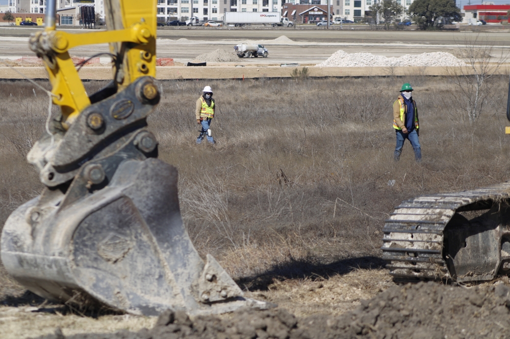 The bottom of a yellow excavator stand in the foreground with two construction works walking through an open site in neon yellow vests.