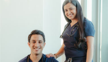 A woman in navy scrubs stands with her hand on the shoulder of a man in navy scrubs sitting in front of her