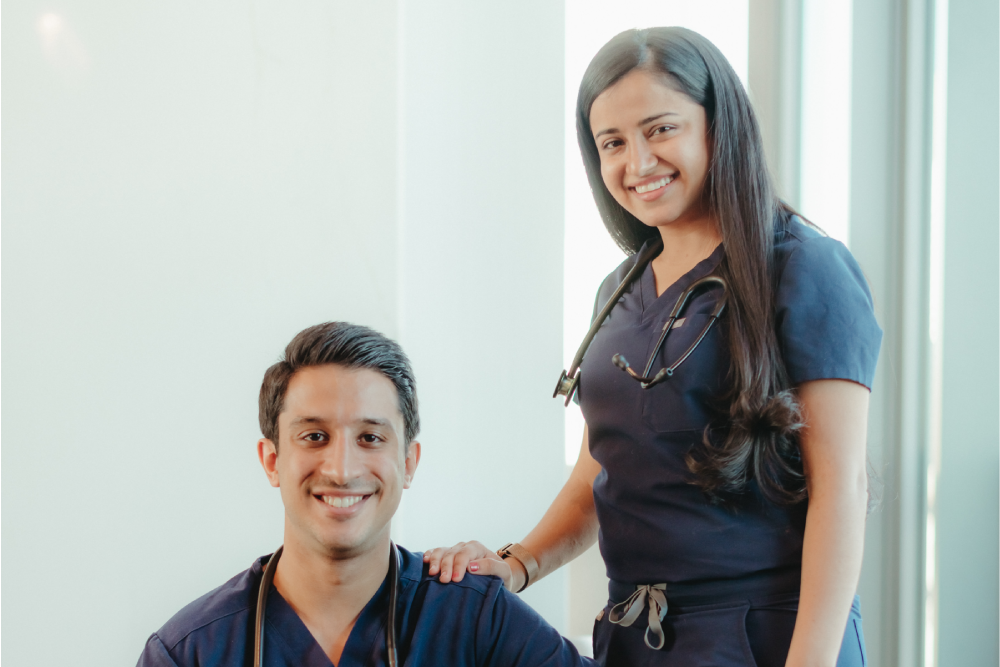 A woman in navy scrubs stands with her hand on the shoulder of a man in navy scrubs sitting in front of her