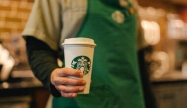 A person wearing a Starbuck's branded green apron holds a lidded paper cup featuring Starbucks branding.