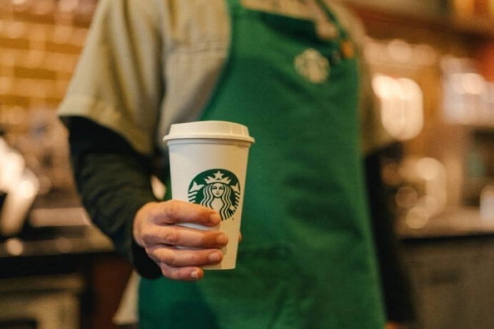 A person wearing a Starbuck's branded green apron holds a lidded paper cup featuring Starbucks branding.