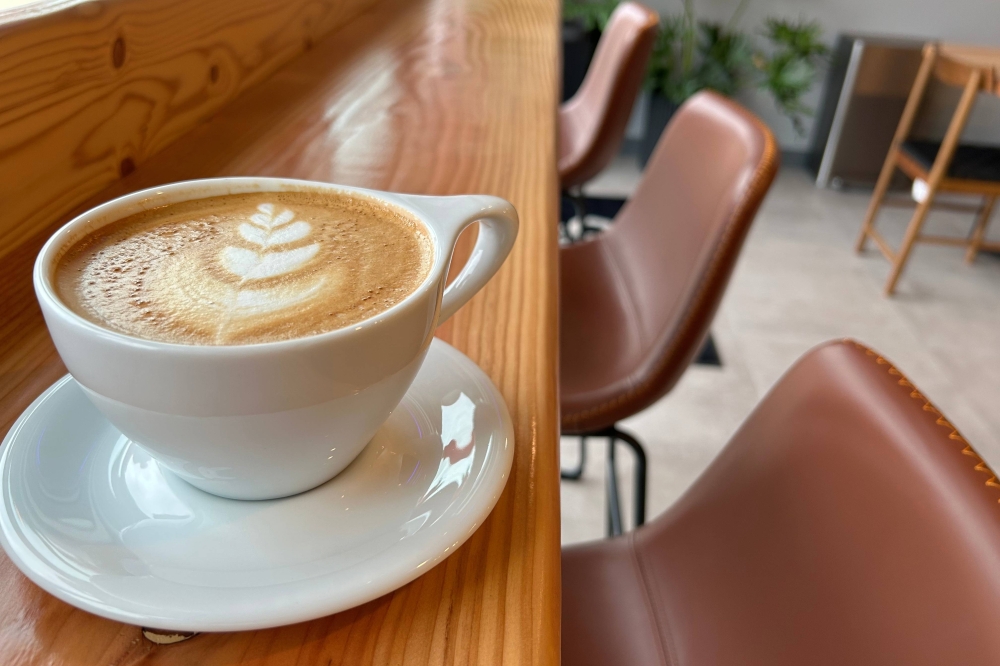 Coffee featuring leaf foam art in a white ceramic cup sitting in a white sauces on a brown wooden bar in a cafe.
