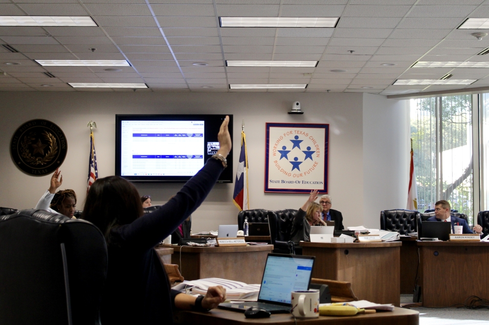 State Board of Education members, seated at desks in a circle, raise their hands to vote on a proposal.