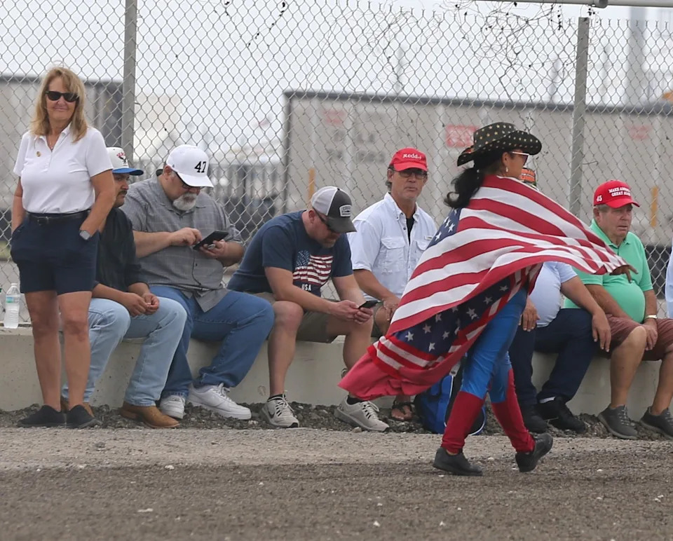 A woman draped in an American flag passes a line of supporters of President Donald Trump outside the Ortiz Center, where Trump was expected to make remarks the afternoon of Feb. 27, 2026.