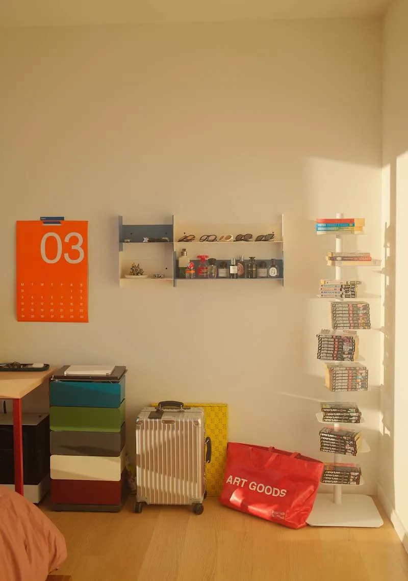 Colorful calendar on the wall, shelves with sunglasses and bottles, a stack of boxes, a suitcase, and a red tote bag.