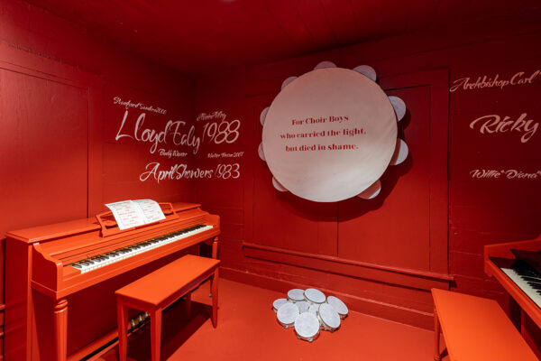 An installation image of a red room with white text and a large white tambourine with text that reads: "For Choir Boys who carried the light, but died in shame." 
