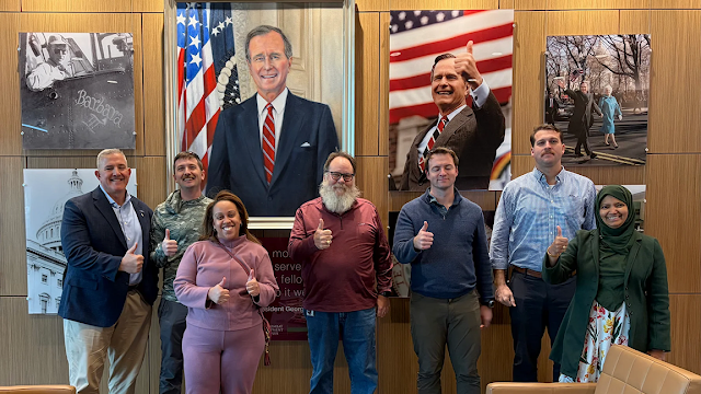 Seven people stand indoors in front of a large framed portrait of former President George H.W. Bush, with several individuals giving a thumbs-up gesture.