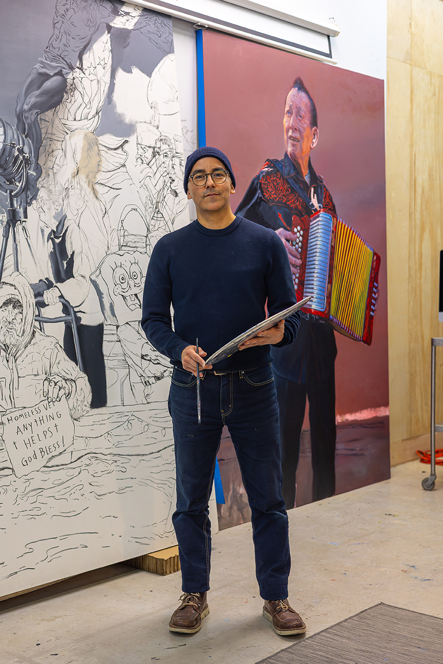 A photograph of artist Vincent Valdez standing in his studio with a palette and paintbrush. Two large-scale paintings are behind the artist.