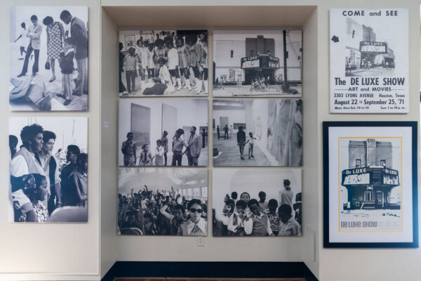 An installation of black and white photograph of the historic "DeLuxe" show featuring people walking through the exhibition.