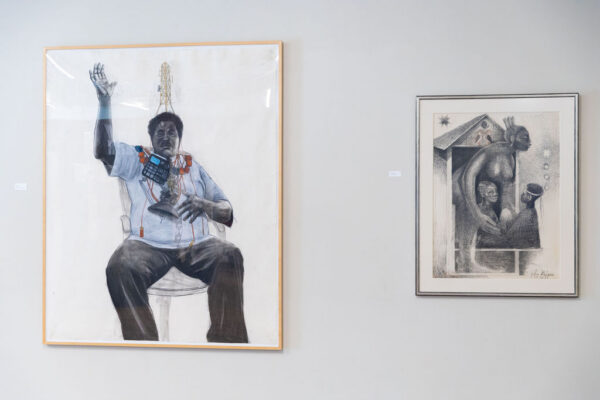 An installation image of a drawing featuring a man covered in communication devices, with his hand raised and a print featuring a woman protecting two children in the doorway of a house.