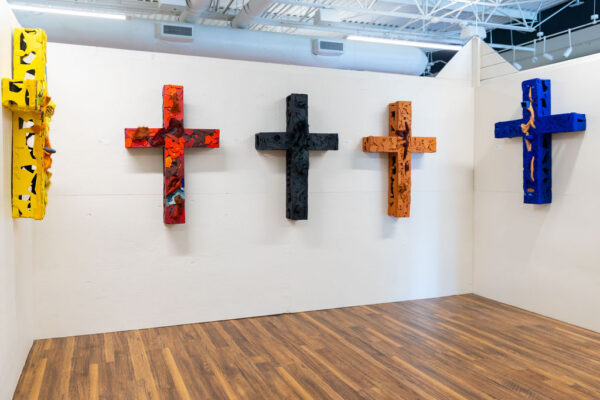 An installation image of five colorful sculptures of crosses hanging on a white gallery wall.