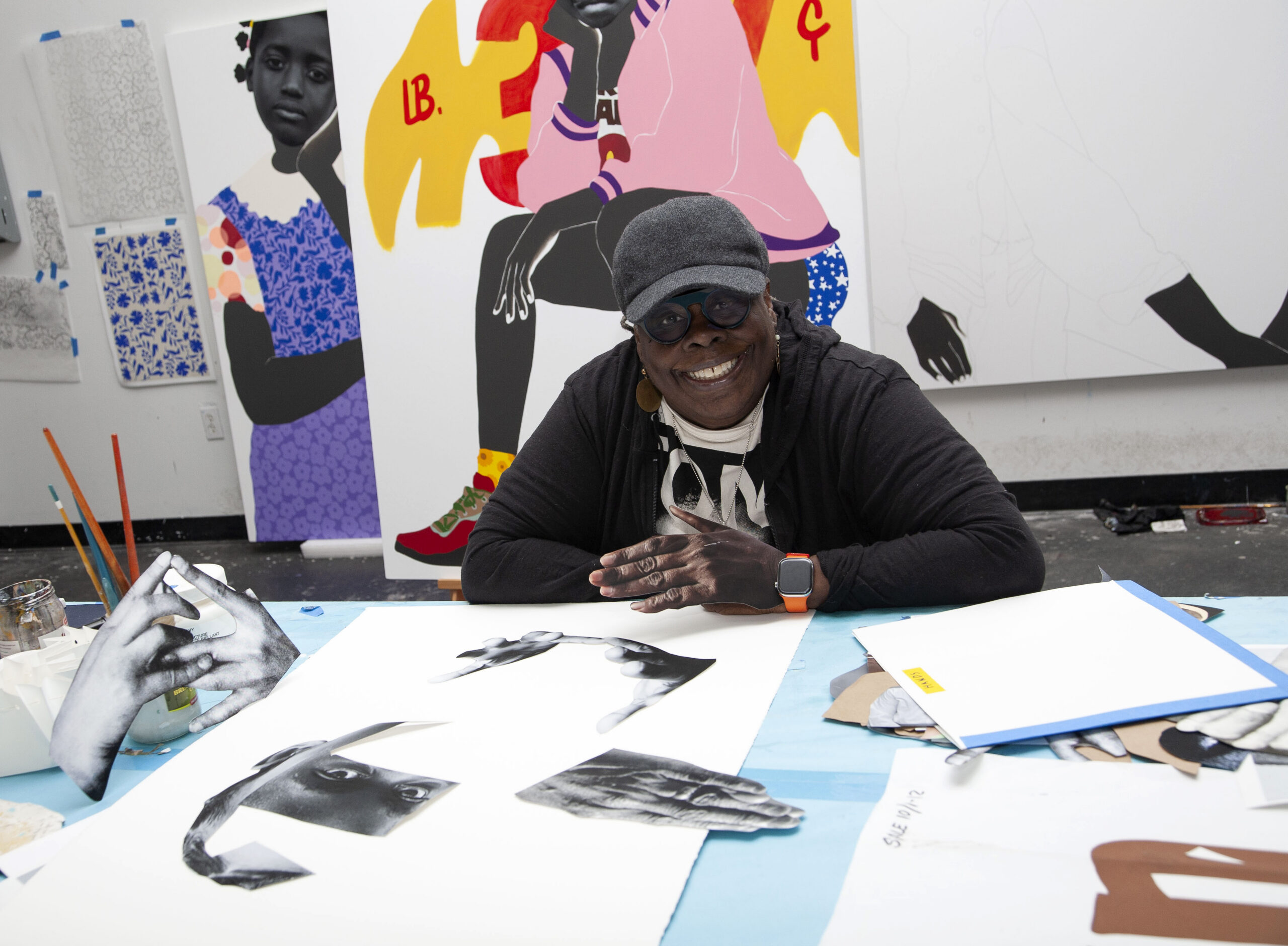 A photograph of artist Deborah Roberts in her studio with cut paper images of a figure on the desk in front of her and a cut paper hand on top of her hand.