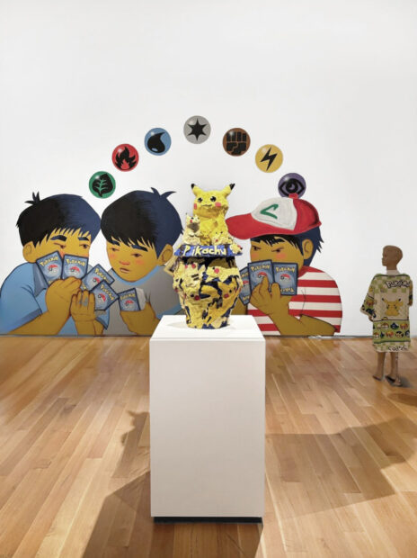 An installation image of a ceramic Pikachu sculpture on a white pedestal in front of a wall mural of children playing with Pokémon cards.