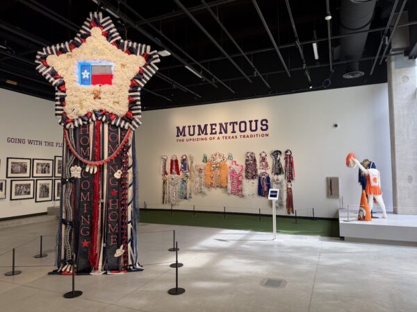 A view of a gallery exhibition titled "Mumentous" with a grouping of framed photographs on one wall, elaborately decorated ribbons on another wall, a cheerldear mannequin, and a large suspended ribbon with lighted Texas flag and the words "HOMECOMING" and "COMING HOME."