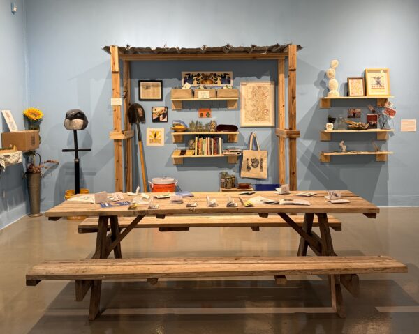 An installation view of an exhibition with a picnic table, bookshelves, farming implements, and artworks.