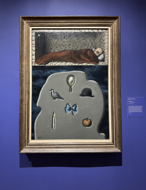 A surrealist painting by René Magritte of a person sleeping with a pillow and blanket in a wooden box. Below the figure is a tombstonelike gray mass imprinted with a bird, mirror, bow, hat, apple, and candle.