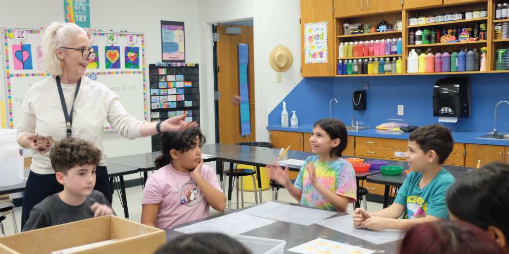 Students smile as an art teacher tosses balls of clay to them around the table.