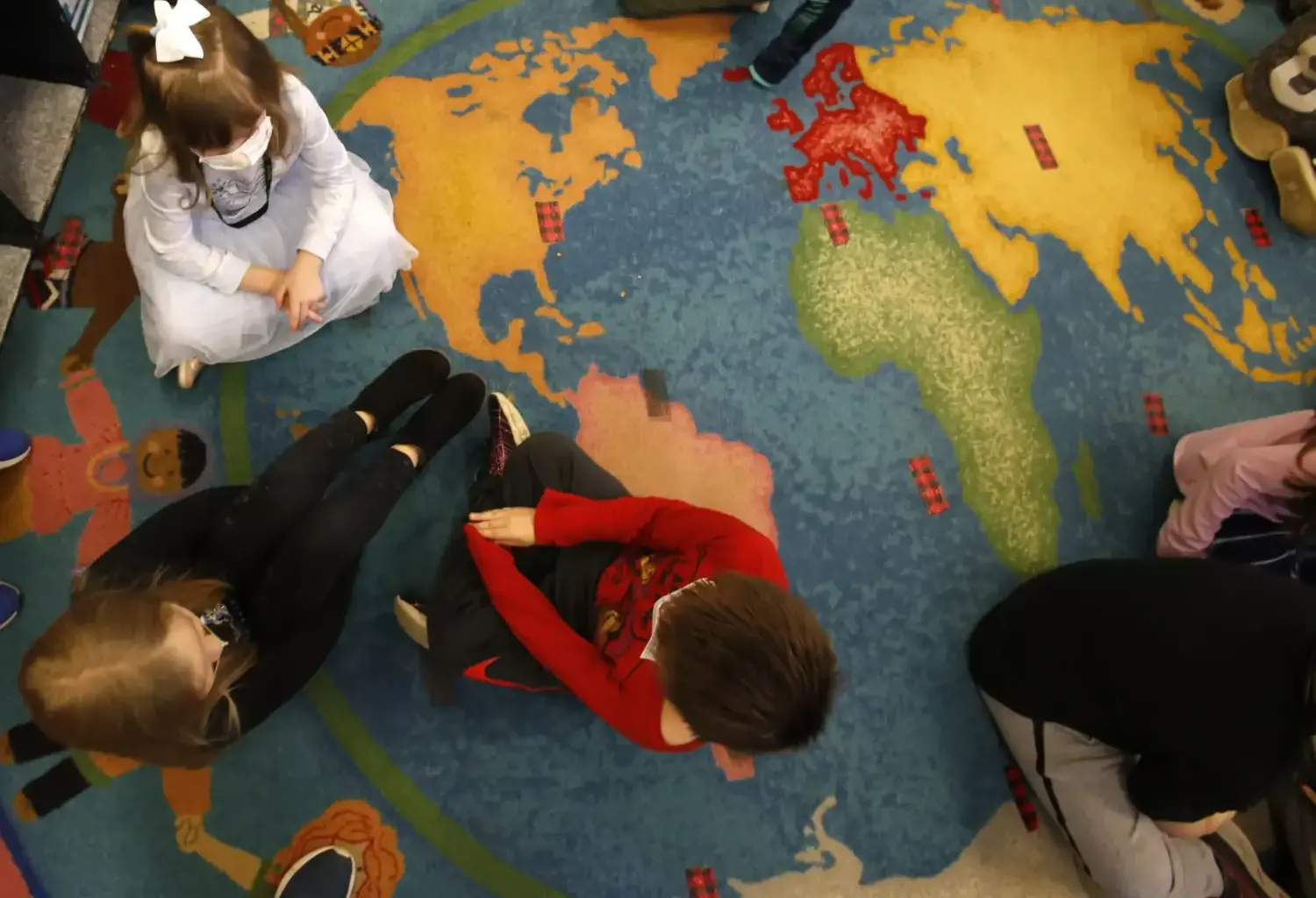 An overhead view shows young children interacting as they sit on a rug that looks like a world map.