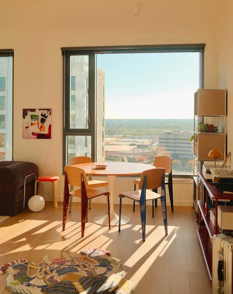 Bright dining area with a round table, colorful chairs, a patterned rug, and large windows showcasing a city view.