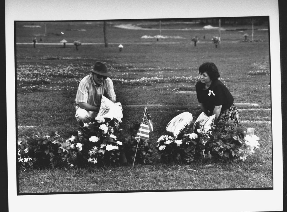 a man and a woman kneel on the ground behind a memorial site filled with flower arrangements, balloons, and a mini american flag