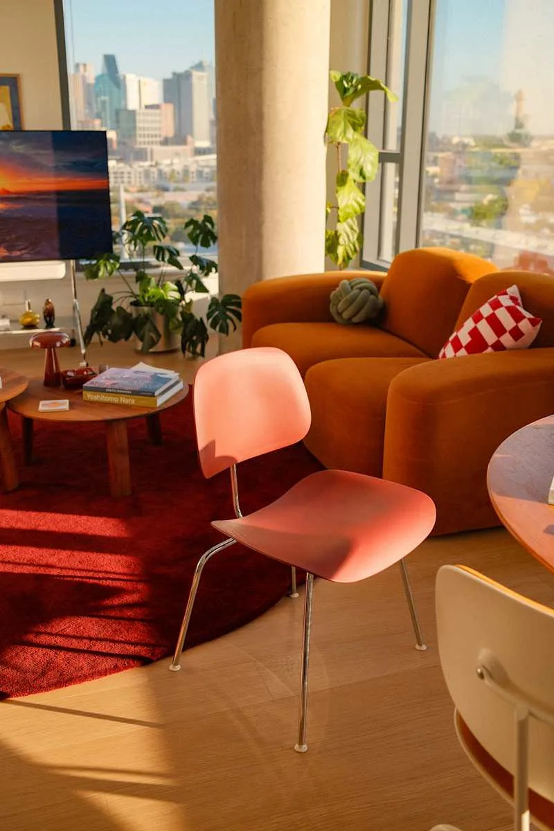 Modern living room with a pink chair, orange sofa, round wooden coffee table, and city skyline view.