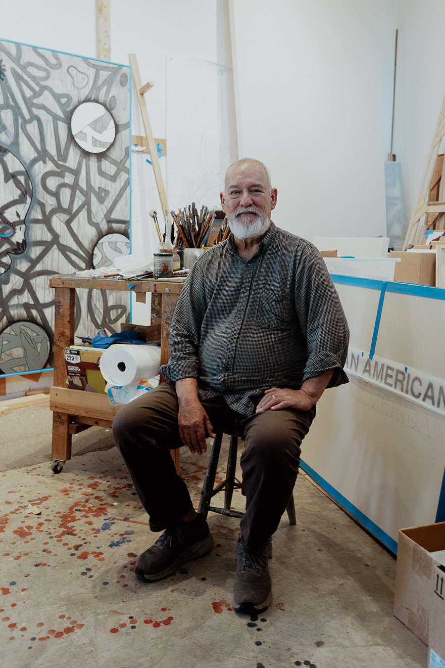 A photograph of artist Benito Huerta, sitting in a chair in his studio with works in progress and materials in the background.