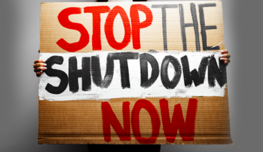A photo illustration of a person holding a cardboard sign with the words "Stop the Shutdown Now."