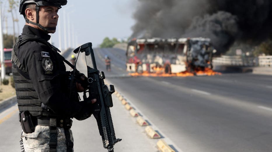 A member of the Prosecutor's Office stands guard near a burning bus at one of the main avenues after it was set on fire by organized crime groups in response to an operation in Jalisco to arrest a high-priority security target in Zapopan, state of Jalisco, Mexico, on February 22, 2026.