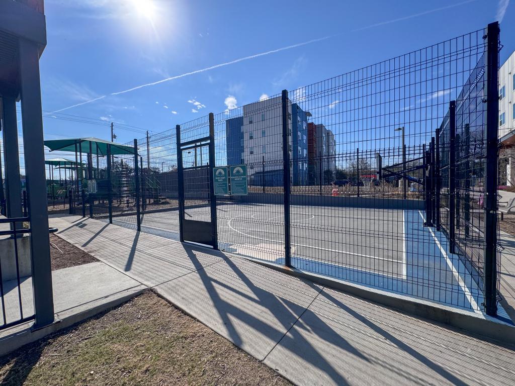 Fenced outdoor sport court with a basketball hoop at Juniper Creek, located beside the playground and residential buildings and designed as an on-site recreation amenity for residents.