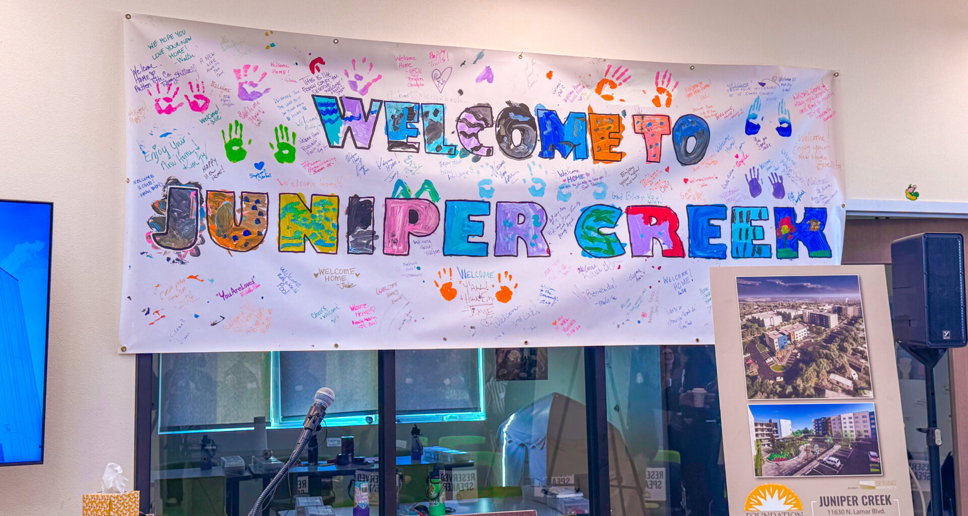 “Welcome to Juniper Creek” banner decorated with colorful handprints and handwritten messages from residents, displayed above a podium and microphone during the Juniper Creek grand opening event. An easel sign shows renderings of the 110-unit affordable housing development.