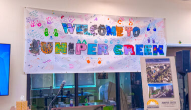 “Welcome to Juniper Creek” banner decorated with colorful handprints and handwritten messages from residents, displayed above a podium and microphone during the Juniper Creek grand opening event. An easel sign shows renderings of the 110-unit affordable housing development.