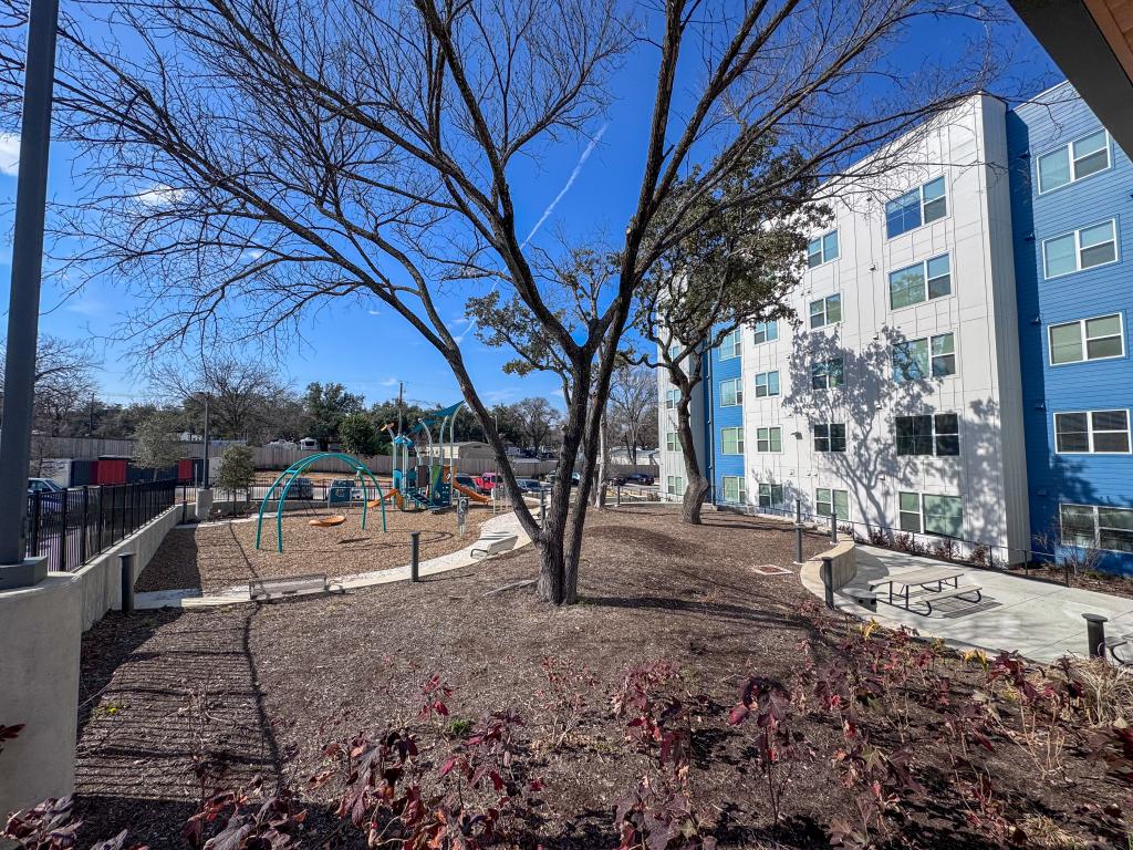 Children’s playground and landscaped courtyard at Juniper Creek, featuring slides, climbing structures, picnic tables, and mature shade trees next to a four-story affordable housing community.