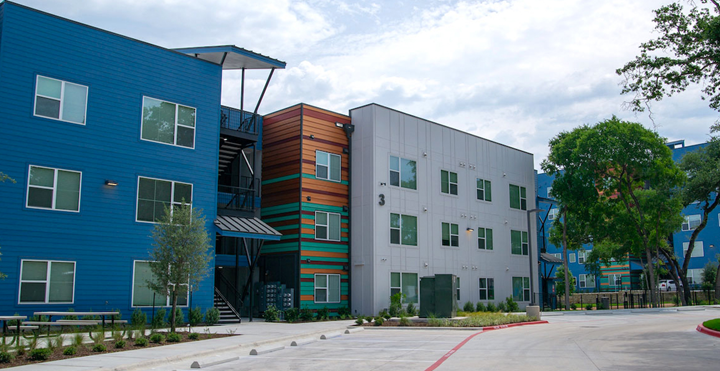 Juniper Creek, a colorful building with the number 3. Blue building on the left with a staircase in between it next to a striped building which is next to a gray building. Surrounded by trees