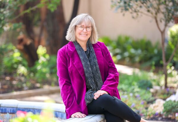 An outdoor portrait of Elizabeth Kathleen Mitchell, smilling in a bright purple jacket and patterned scarf.