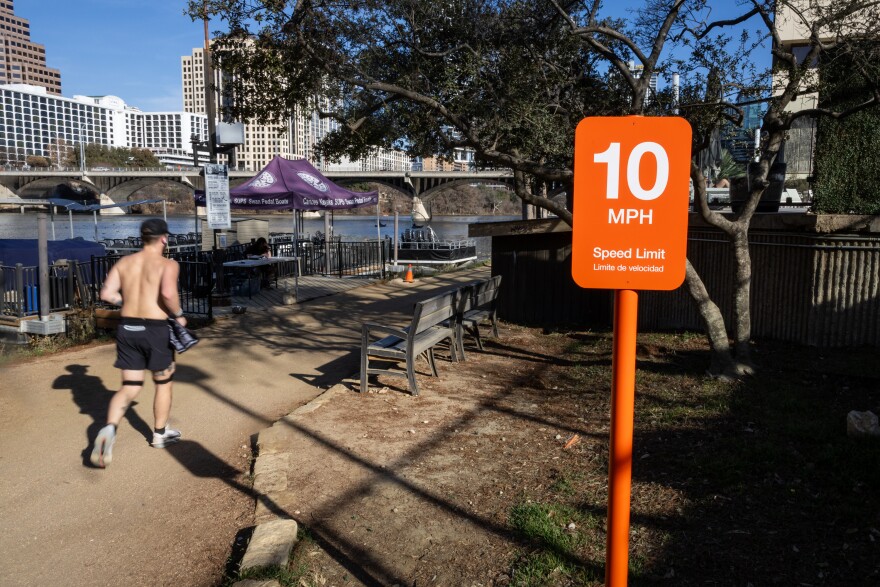  A person runs on a trail along a river with the city in the background. Also shown is an orange sign that reads 10mph.