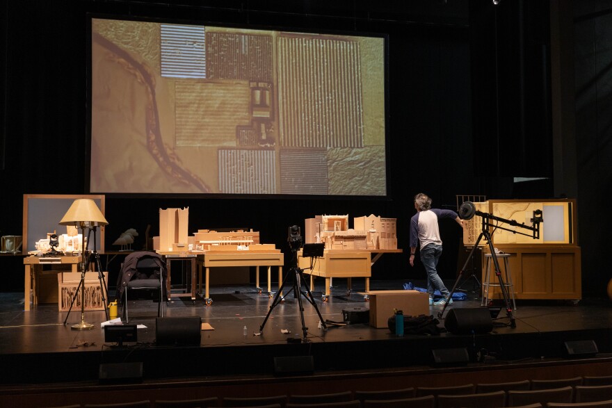 A man stands onstage amongst an art project with a projection. 
