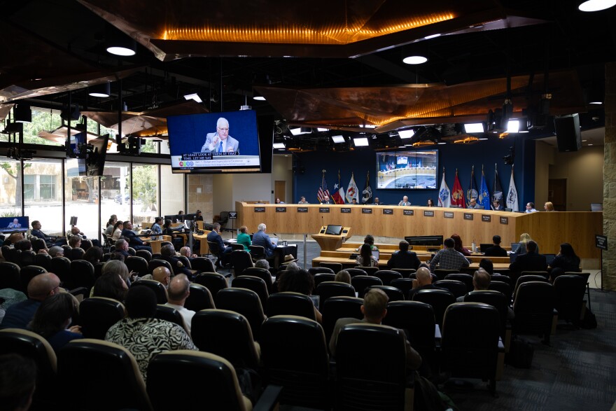 Austin City Council chambers. The audience is in the foreground with the council dais in the background. On a TV screen, Mayor Kirk Watson is seen speaking. 