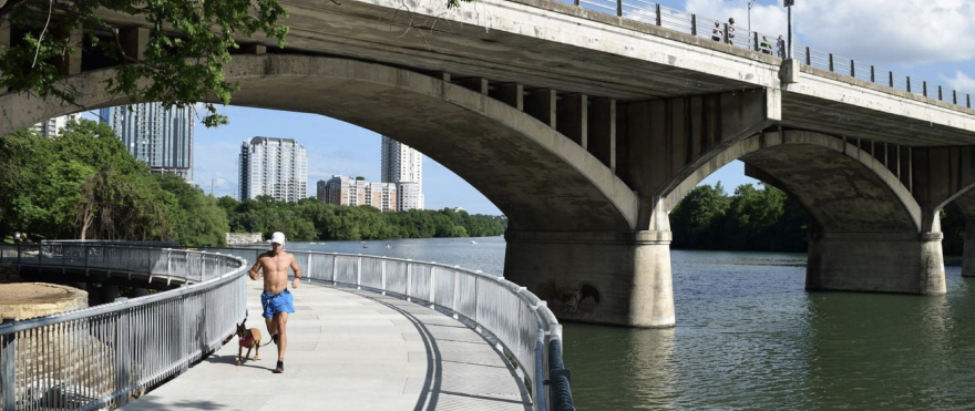 A shirtless man is running with a dog on a trail bridge beneath the Congress Avenue Bridge.