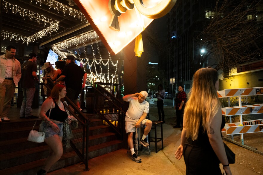 A man sits on a stool outside of a bar as people pass. He has his arm up near his ear.