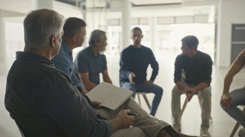 Professional psychologist taking notes in a group therapy session in an office. Diverse men with mental health issues talk about their feelings and emotions while supporting one another in recovery