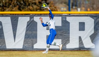 A softball player wearing an SDSU uniform and blue helmet stands on the field holding a ball, with the outfield fence and a building visible in the background.